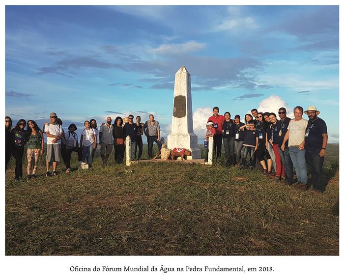 4 z 8- Oficina do Fórum Mundial da Água na Pedra Fundamental, em 2018. Imagem retrata homens, mulheres e crianças posando ao lado da Pedra Fundamental,. Em destaque uma peça artesanal( uma capivara em madeira, com um tecido vermelho e branco por cima), encontra-se aos pés da pedra fundamental. Realização: Academia Planaltinense de Letras, Artes e Ciências (APLAC), pelo Ecomuseu Pedra Fundamental e pelo Coletivo Nativo Audiodescrição produzida  pelo Instituto de Promoção das Pessoas com Deficiência Visual Audiodescritora: Elma Lúcia Rodrigues Consultor: Fernando Rodrigues Este projeto é promovido com recursos do Fundo de Apoio a Cultura do DF